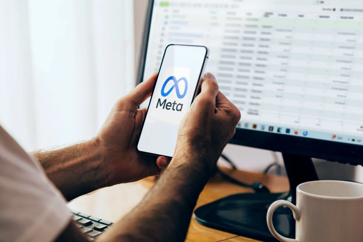 Person holding a smartphone with the Meta logo while reviewing digital advertising campaign data on a computer.